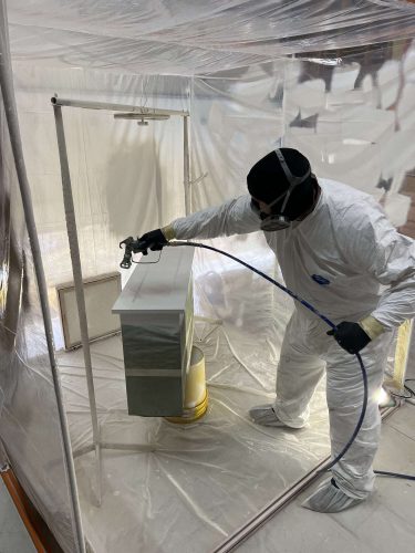 A painter from Pearl Painters in Portland spray paints white cabinets inside a plastic enclosure, prepping them for installation by Evans in April 2025.