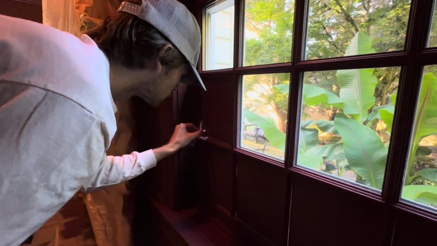 A painter in a cap opens a dark window, showing greenery and outdoor furniture at a Purcell home, Portland, July 2025.