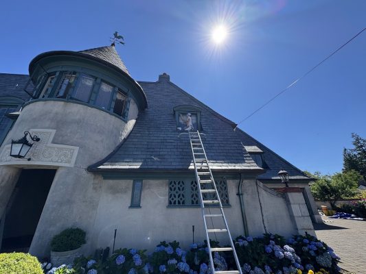 Painter sits at upstairs window of gray stucco house in Portland; ladder against roof, bright August 2025 sun, clear sky.