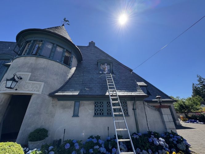 Painter sits at upstairs window of gray stucco house in Portland; ladder against roof, bright August 2025 sun, clear sky.