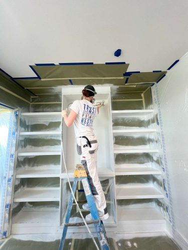 A painter from Pearl Painters in Portland spray-paints built-in cabinets for an Isadore renovation in April 2025. The room is protected with plastic and painter's tape.