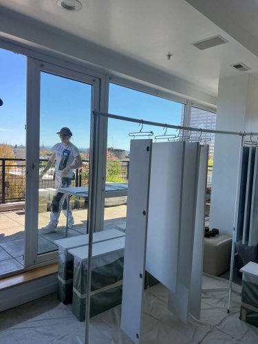 A painter from Pearl Painters is on a balcony outside glass doors in Portland. Inside, cabinets and painted panels are visible, with furniture covered by drop cloths. This Isadore painting project is scheduled for completion in April 2025.