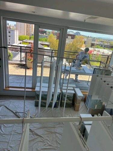A painter from Pearl Painters works with white cabinets on an apartment balcony in Portland, with city buildings and trees in the background. The painting project is happening around April 2025.