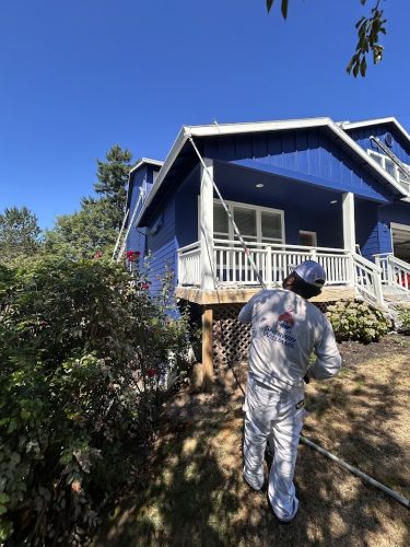 A painter in white uses a pole to paint a blue house gutter in Jennings, July 2025. Pearl Painters, not Portland.