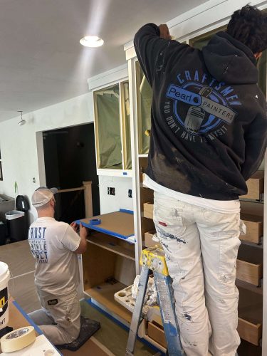 Two Evans painters prepare and paint kitchen cabinets; one tapes a surface while the other paints a cabinet frame from a ladder. This painting work is done by Pearl Painters in Portland.