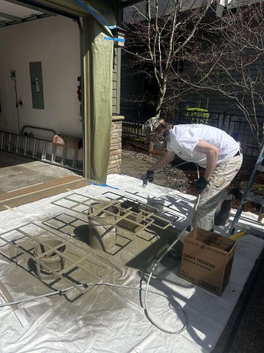In April 2025, painter Taylor spray paints metal grates in a garage set up for exterior painting, using protective gear, paint thinner, and coverings on the floor and walls. This project took place in Portland and was part of Pearl Painters' work.