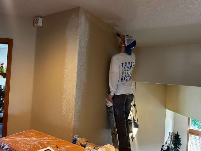 In April 2025, a painter from Pearl Painters paints a high interior wall with a roller in Portland. Furniture is covered with plastic.