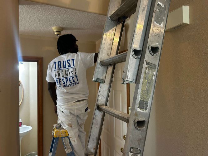 A painter from Pearl Painters works on a hallway ceiling near a door in Portland, standing on a small ladder with a larger ladder visible nearby. Photo taken April 2025.