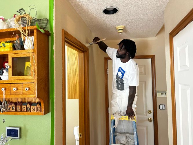 A painter from Mann Interior is on a ladder painting a hallway wall, holding a paint tray and brush. Shelves with decor and a thermostat are nearby. The painting took place in April 2025. Location not specified, but painting services like Pearl Painters are available in Portland.