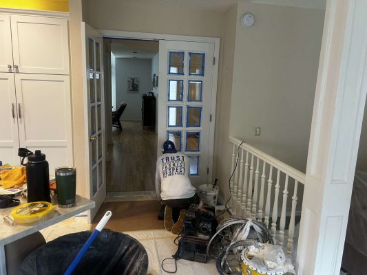 A hallway at Mills in Portland is shown under renovation with painting supplies, taped French doors, a covered floor, and tools by a white railing. Photo captured April 2025 by Pearl Painters.