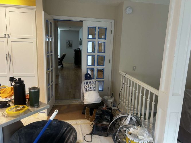 A hallway at Mills in Portland is shown under renovation with painting supplies, taped French doors, a covered floor, and tools by a white railing. Photo captured April 2025 by Pearl Painters.