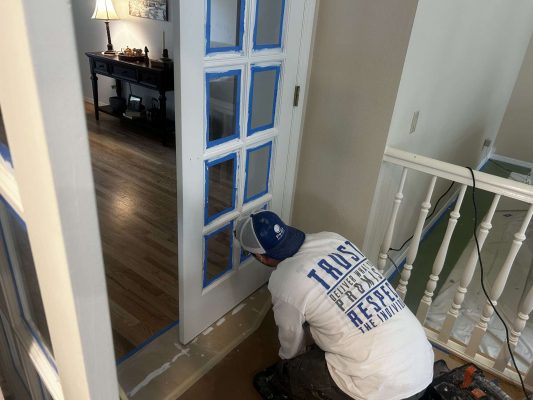 A painter in Mills kneels while painting the interior trim of a glass-paneled door, with blue painter’s tape and supplies visible. (April 2025).