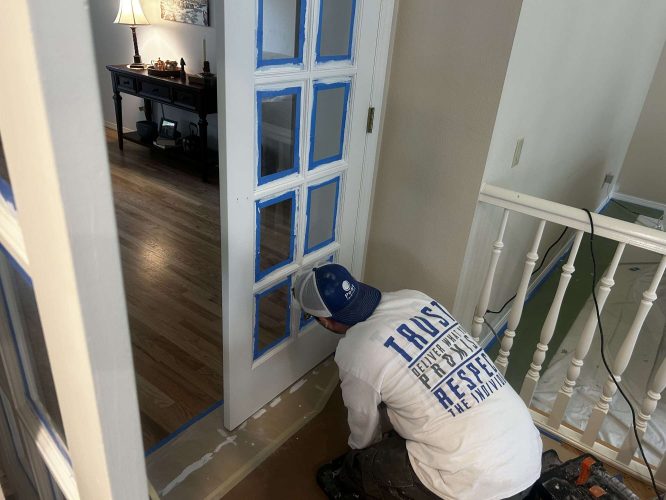 A painter in Mills kneels while painting the interior trim of a glass-paneled door, with blue painter’s tape and supplies visible. (April 2025).