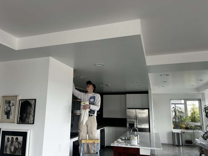 A painter from Pearl Painters stands on a ladder in a Portland kitchen, painting the upper corner of modern white cabinets. Paint cans and framed photos are nearby as the space is prepared for an Isadore gathering in April 2025.