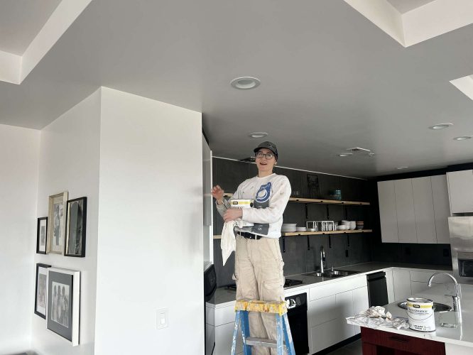 In April 2025, painter Isadore stands on a ladder painting the upper wall of a modern kitchen in Portland. Painting supplies and sleek cabinets are visible nearby. This work is part of a Pearl Painters project.