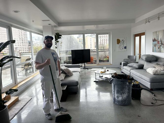 Isadore, a painter with Pearl Painters in Portland, stands with a broom in a modern living room with large windows and grey furniture, preparing the space for painting in April 2025.