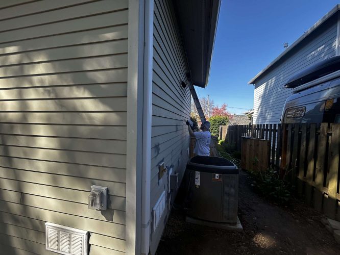 In April 2025, a painter in a white shirt uses a ladder to work near the roof of a beige house exterior beside an outdoor HVAC unit, possibly in the Congdon area. Another house and van are nearby. This may involve Pearl Painters from Portland.