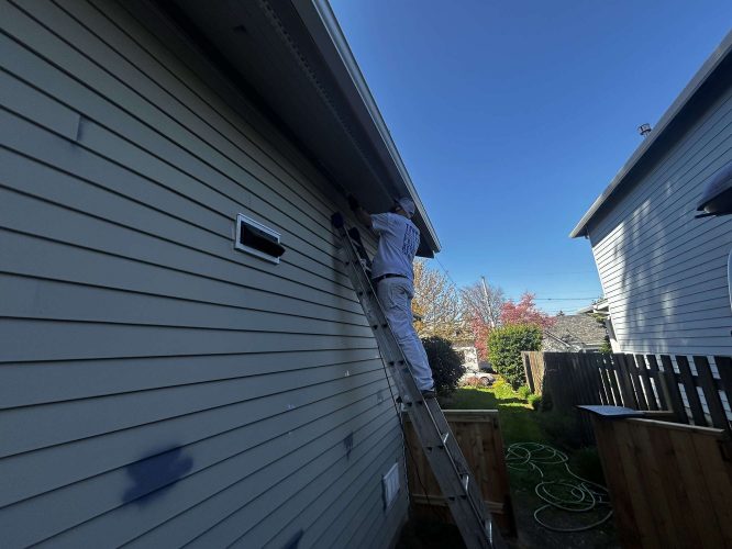 In April 2025, a painter from Pearl Painters paints the exterior trim near the roof of a light-colored Congdon house in Portland on a sunny day.