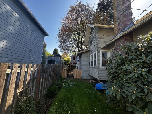 Pearl Painters documented this Portland exterior painting project in April 2025: a beige house with a brick chimney, fenced backyard, green grass, large shrub, and nearby Airstream trailer.