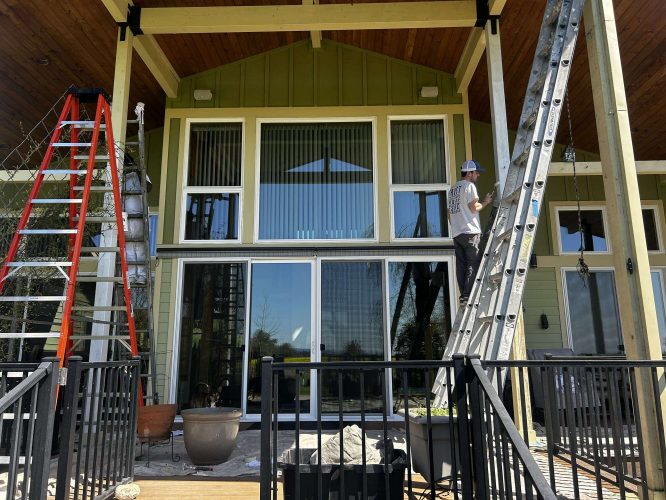 A painter from Pearl Painters works on the exterior of a two-story house with large windows and a covered porch in Portland as part of the McCaffery painting project, scheduled for completion in April 2025.