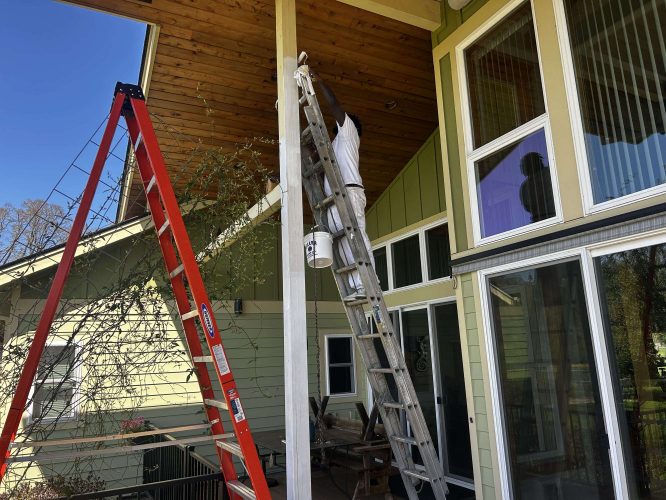 In April 2025, a painter in Portland paints an exterior vertical beam of a house while standing on a tall ladder with a paint bucket. A red step ladder nearby is part of the McCaffery renovation project by Pearl Painters.