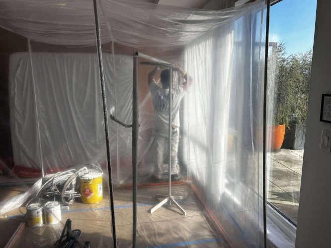 A painter in white coveralls works on cabinets inside a plastic-sheeted area near a large glass door. Painting supplies are on the floor, indicating Pearl Painters is preparing for Isadore’s April 2025 renovation project in Portland.