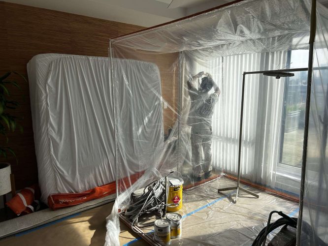 A painter from Pearl Painters is inside a plastic enclosure at an Isadore project in Portland, with painting supplies and covered furniture nearby. Completion is expected by April 2025.