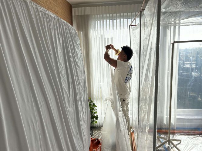 A painter from Pearl Painters in Portland tapes clear plastic to a window and covers the workspace with protective sheets, preparing for cabinet painting in April 2025.