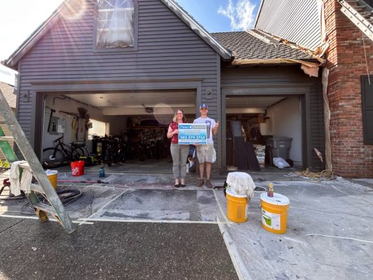 Renee & Seamus Barron hold a Pearl Painters sign by a garage under repair in Portland, with tools and materials around.