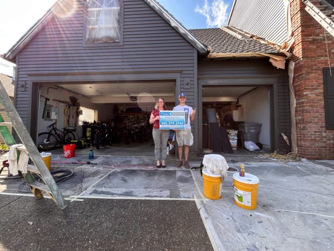 Renee & Seamus Barron hold a Pearl Painters sign by a garage under repair in Portland, with tools and materials around.