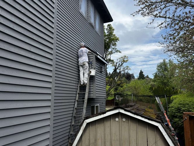 Painter Scott from Pearl Painters paints the upper exterior siding of a gray house next to a shed in Portland, with trees and power lines visible in April 2025.