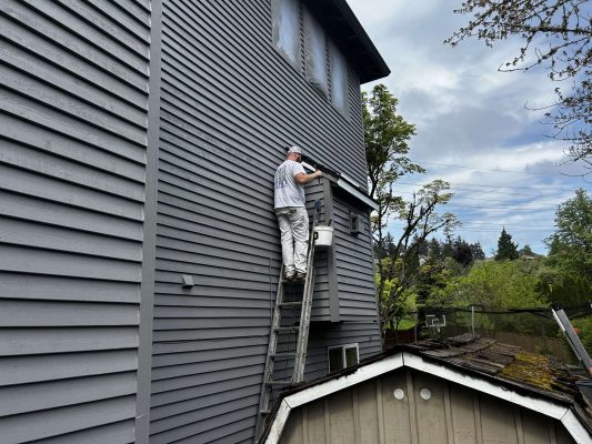 In April 2025 in Portland, painter Scott from Pearl Painters paints the exterior of a tall gray house above a small shed, with trees and power lines behind him.