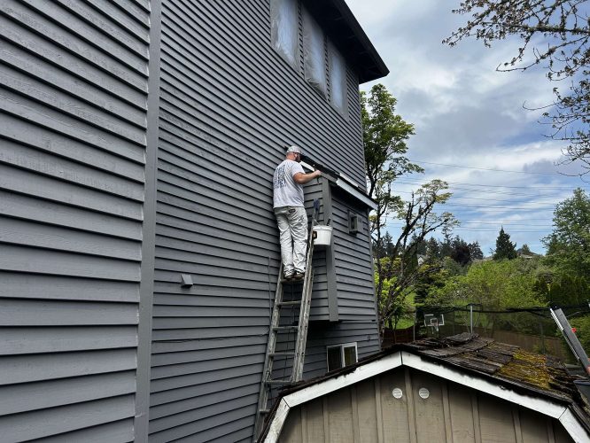 In April 2025 in Portland, painter Scott from Pearl Painters paints the exterior of a tall gray house above a small shed, with trees and power lines behind him.