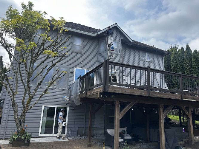 In April 2025, two painters from Pearl Painters paint the exterior of a gray house in Portland; Scott works on a ladder near the roof while another painter works by the sliding glass door at ground level.