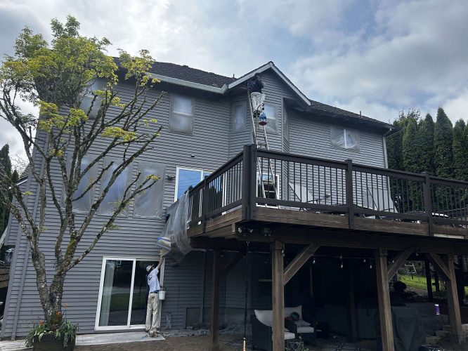 In April 2025, Pearl Painters workers prepare a gray two-story Portland house for painting by covering the windows and deck with plastic sheets. Painter Scott works on a ladder while another painter assists from the ground.