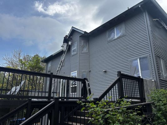 In April 2025, painter Scott from Pearl Painters paints the upper exterior of a gray two-story house in Portland, using plastic window covers near a black wooden deck.