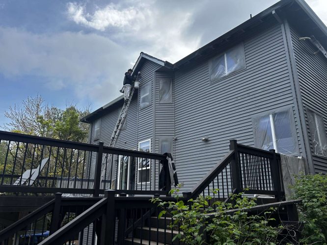 In April 2025, painter Scott from Pearl Painters paints the upper exterior of a gray two-story house in Portland, using plastic window covers near a black wooden deck.