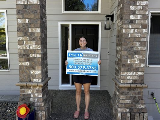 Person in front of Featherstone house holds Pearl Painters sign for Portland painting services, available through Aug 2025.