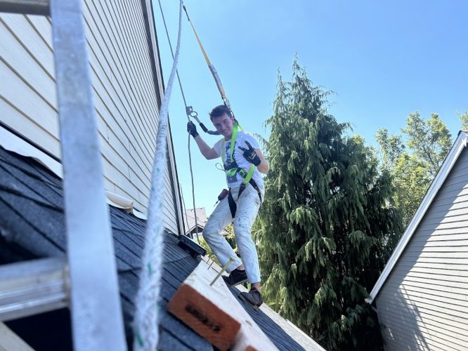 A painter in safety gear stands on a sloped roof in Portland, secured by harness; painting by Pearl Painters, August 2025.