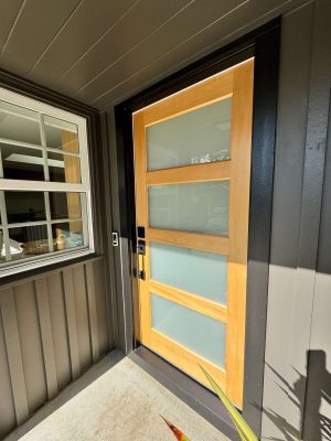 Modern Mastalir exterior wooden front door with three frosted glass panels, black frame, and metal handle on a covered porch in Portland. Pearl Painters recommends this style for May 2025 painting projects.