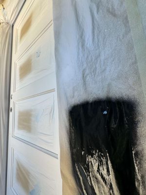 Pearl Painters, a Portland painter, is spray painting a door for a Mastalir project. The door has some white areas and one large black exterior section, with protective tape and plastic applied. Completion is scheduled for May 2025.
