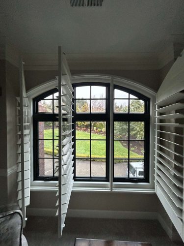 A large interior window with black grid panes and open white shutters at McCormick provides a view of the landscaped garden and lawn. Pearl Painters, a Portland painting company, highlights this peaceful setting in April 2025.