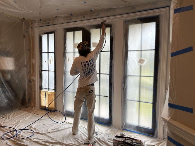 In April 2025, a painter with Pearl Painters in Portland is spray painting interior window frames in a room protected with plastic sheeting during renovation.