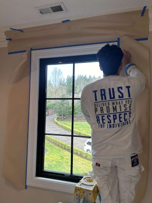A painter from Pearl Painters prepares an interior window for painting by taping brown paper around it. The photo was taken at McCormick in April 2025, outside Portland. Their jacket features the words, "TRUST, DELIVER WHAT YOU PROMISE, RESPECT THE INDIVIDUAL.