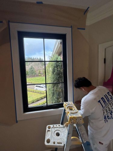 A painter from Pearl Painters in Portland is shown in April 2025 at McCormick, standing on a ladder and taping around a window frame with a street view outside.