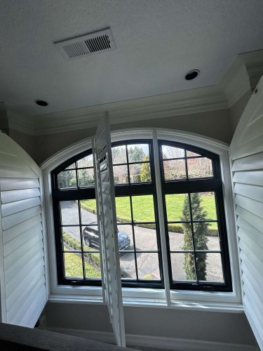 A painting by Pearl Painters in Portland shows a view through an arched window with black trim and white shutters, overlooking a driveway, black car, and grassy yard with trees in April 2025.