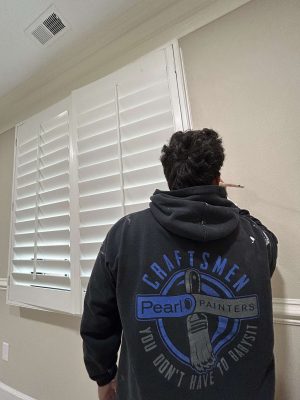 A painter in a "Craftsmen Pearl Painters" hoodie paints the edge of a beige wall near white shutters, demonstrating painting skills suitable for an April 2025 McCormick home refresh.