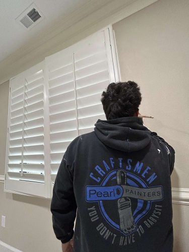 A painter in a "Craftsmen Pearl Painters" hoodie paints the edge of a beige wall near white shutters, demonstrating painting skills suitable for an April 2025 McCormick home refresh.