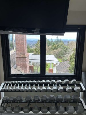 In April 2025, Pearl Painters photographed a McCormick interior in Portland featuring a rack of dumbbells by a window with views of trees, rooftops, and a brick pillar; a TV is mounted above.