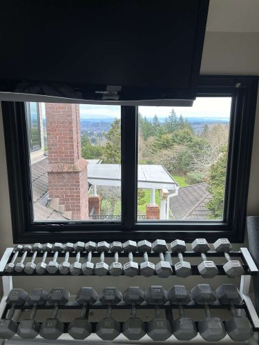 In April 2025, Pearl Painters photographed a McCormick interior in Portland featuring a rack of dumbbells by a window with views of trees, rooftops, and a brick pillar; a TV is mounted above.